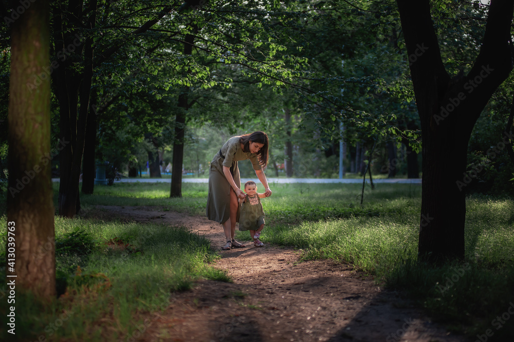 Fototapeta premium The baby girl takes the first steps, a young mother holding a child by the hands, insuring, helping to walk along the path of the park amid green trees and grass. Parental care, walks in the fresh air