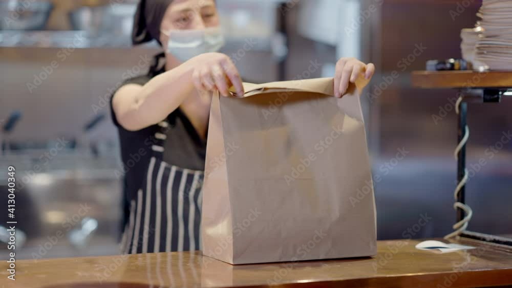 Blurred Caucasian young woman in Covid face mask packing takeaway food in cafe or restaurant. Professional cook preparing lunch order in slow motion indoors on coronavirus pandemic. Blurred Caucasian young woman in Covid face mask packing takeaway food in cafe or restaurant. Professional cook preparing lunch order in slow motion indoors on coronavirus pandemic.