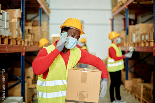Afro male worker carrying a box and talking on the phone in a warehouse. Focus on faces