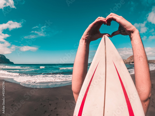 Surfer heart shaped hand gesture from behind the surfboard on the black sand beach background.