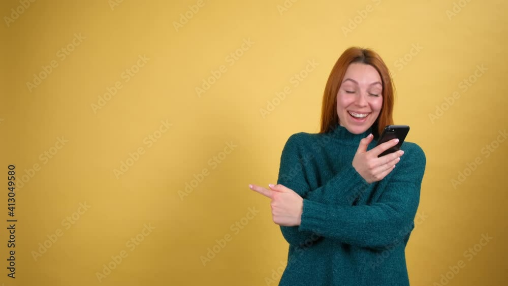 Young fun woman 20s in green sweater say wow hold in hand using mobile cell phone pointing fingers aside on workspace. Copy space isolated on yellow background in studio. People lifestyle concept Young fun woman 20s in green sweater say wow hold in hand using mobile cell phone pointing fingers aside on workspace. Copy space isolated on yellow background in studio. People lifestyle concept