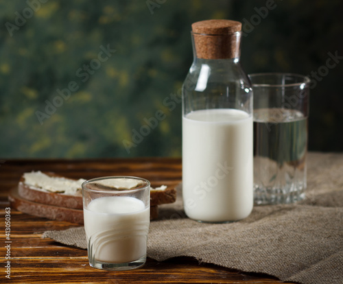 A glass of milk, a glass bottle of milk on the back, and a piece of rye bread and butter on a wooden background.
