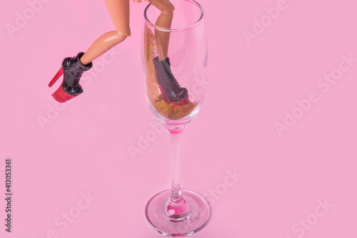 Minimal composition with female doll with trendy  high heels walking and falling into champagne glass of gold glitter on pink background