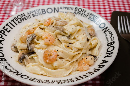 Delicious fettuccine with cream sauce of mushrooms and shrimp on a plate that says Bon Appetite with red checkered tablecloth.