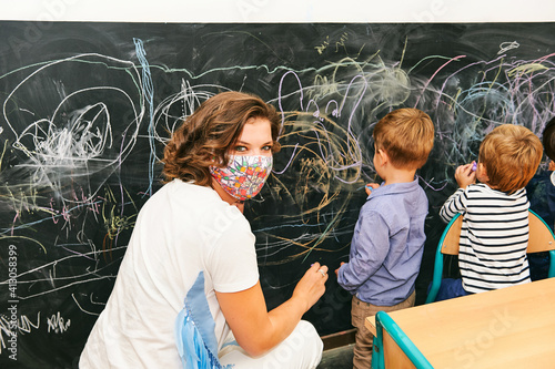 Art classroom in kindergarten, young teacher drawing with children on chalk wall, wearing facemask