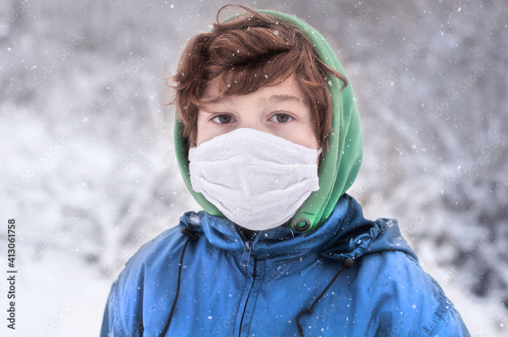 Portrait of a poor boy in a reusable protective mask in the winter outdoors. Pandemic coronavirus. Quarantine covid-19. Winter holidays in lockdown. Snowy weather. Boy in a hood without a hat in frost