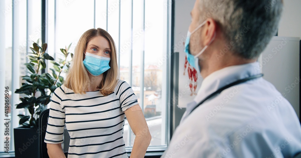 Fototapeta premium Rear of Caucasian senior experienced handsome professional male physician specialist sitting in clinic cabinet making injection to pretty female patient in medical mask vaccination coronavirus concept