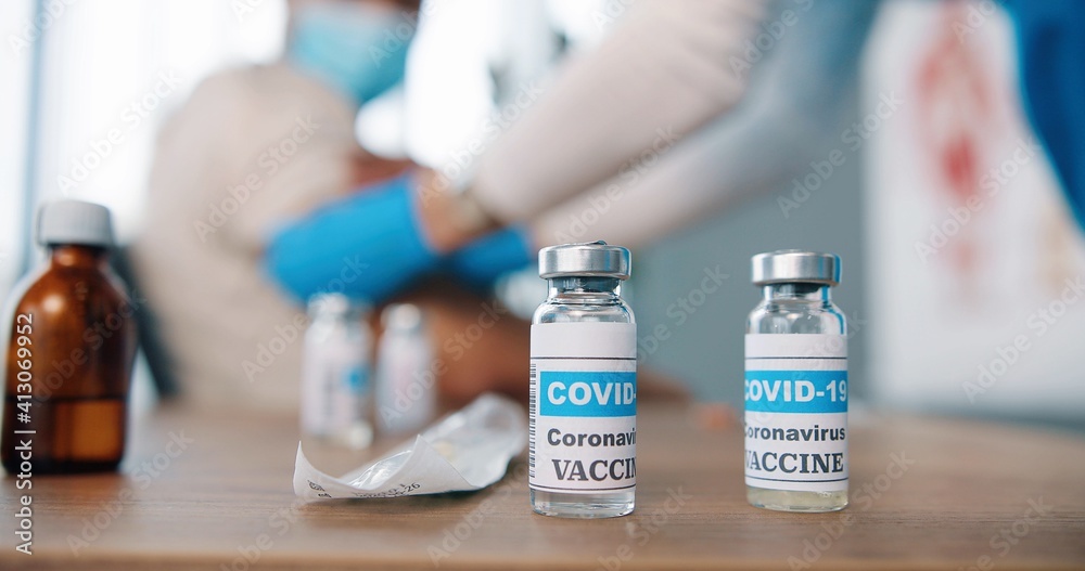 Close up shot of coronavirus vaccine ampoules with liquid, ampoule with vaccine from covid-19 on table in clinic room, doctor making injection on background, injection set, vaccination concept