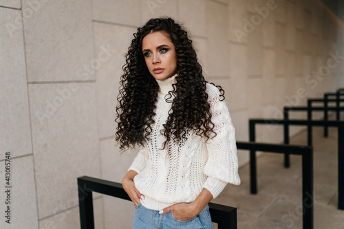 A girl with a sad expression poses in a white sweater and blue jeans with her hands in the front pockets of her trousers. High quality photo