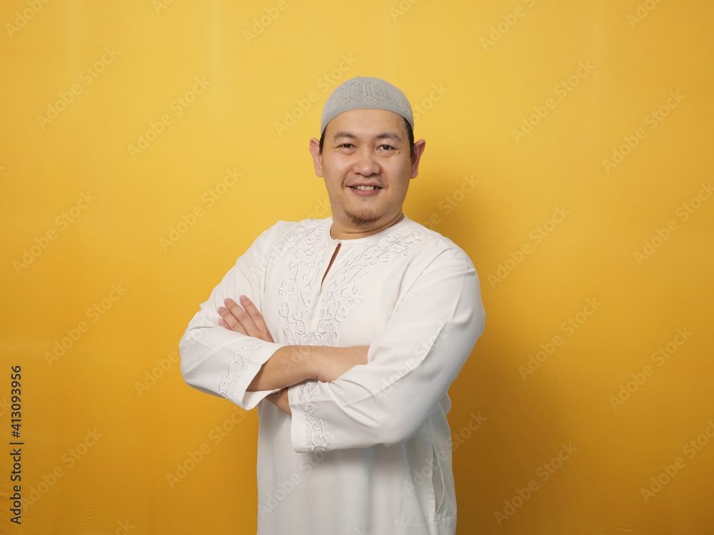 Portrait of Asian muslim man smiling friendly with crossed arms, confident gesture, successful businessman, against yellow