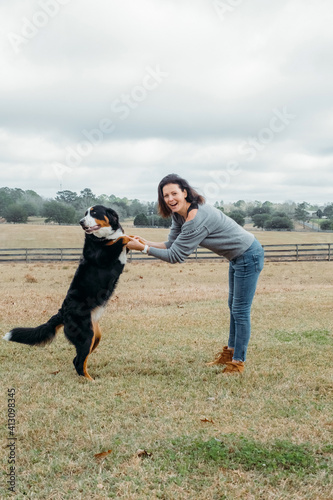 Happy woman with active dog playing outdoor. Cheerful owner and big bernese mountain dog have fun on field against village landscape. Walking with pet.