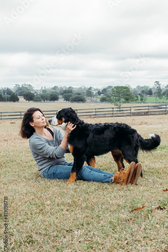 Happy woman with active dog playing outdoor. Cheerful owner and big bernese mountain dog have fun on field against village landscape. Walking with pet.