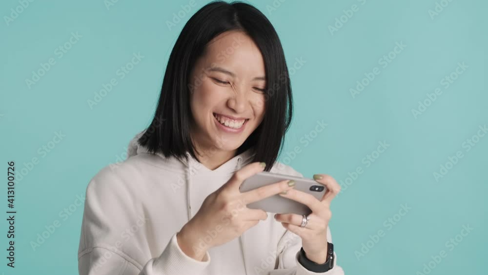 Excited Asian girl emotionally playing on smartphone being obsessed with online games over blue background. Modern technology Excited Asian girl emotionally playing on smartphone being obsessed with online games over blue background. Modern technology