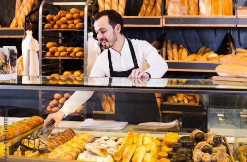 happy male shop assistant demonstrating fresh delicious pastry in bakery