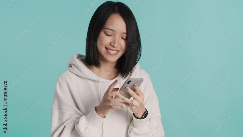 Pretty smiling Asian girl in white hoodie chatting with boyfriends online on smartphone looking cute isolated on blue background. Modern technology Pretty smiling Asian girl in white hoodie chatting with boyfriends online on smartphone looking cute isolated on blue background. Modern technology