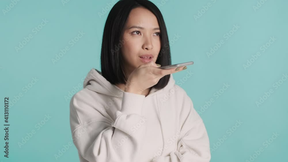 Attractive Asian girl recording voice message to friend using smartphone isolated on blue background. Distant communication Attractive Asian girl recording voice message to friend using smartphone isolated on blue background. Distant communication