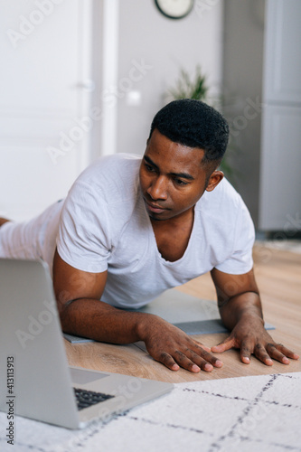 Close-up view of young African-American man exercising workout in horizontal plank position using laptop to watching yoga and training video digital tutorial.