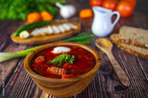 Borscht with sour cream in a wooden bowl on a wooden table. The table also contains black bread, bacon, green onions, peppers and garlic. Traditional Ukrainian and Russian cuisine.