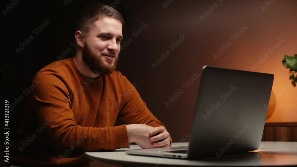 Close-up of smiling young man chatting online on laptop camera at dark living room. Portrait of joyful bearded male making video call at home. Handsome guy gesturing with hand in front of web camera. Close-up of smiling young man chatting online on laptop camera at dark living room. Portrait of joyful bearded male making video call at home. Handsome guy gesturing with hand in front of web camera.
