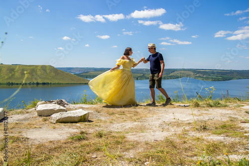 A pretty girl in a long yellow dress dancing with guy on a high cliff by the bay or river on sunny day. Young beautiful woman with short hair having fun with man on the shores of the big river © pijav4uk