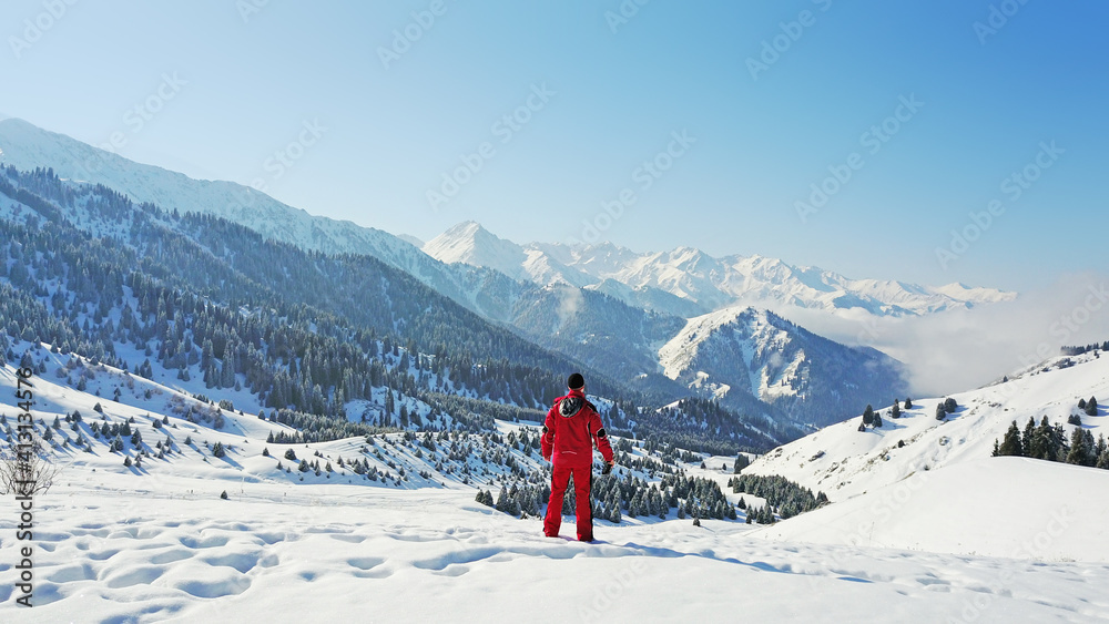 The guy looks at the snow hills and clouds. Mountainous terrain, all covered with fresh snow. Blue sky and huge clouds float along the gorge. On the slopes of a small forest, coniferous trees.