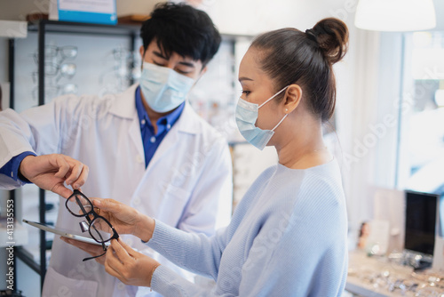 Asian optometrist in white coat and medical mask gives senior woman advice on new model of glasses