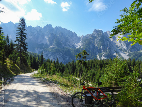 An orange mountain bike leaning against a bench next to a gravelled road in the mountains with the view on high Alps in the region of Gosau, Austria. The chains in the back are stony and barren. Calm