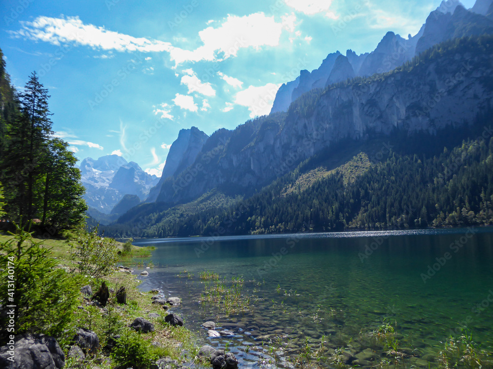 Panoramic view on Gosau lake, with Dachstein glacier in the back in Austrian Alps. The lake is surrounded by high mountains, overgrown with tall trees. Sun reflects on the surface. Serenity  and calm