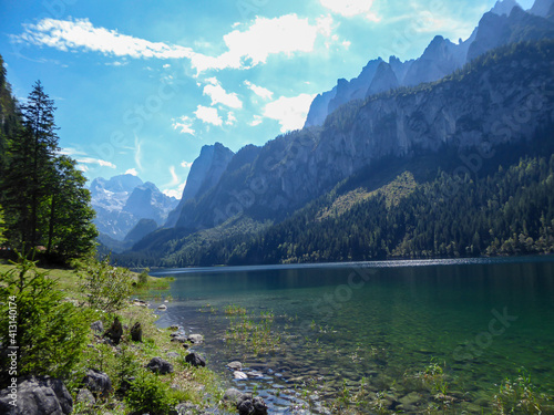 Panoramic view on Gosau lake, with Dachstein glacier in the back in Austrian Alps. The lake is surrounded by high mountains, overgrown with tall trees. Sun reflects on the surface. Serenity and calm