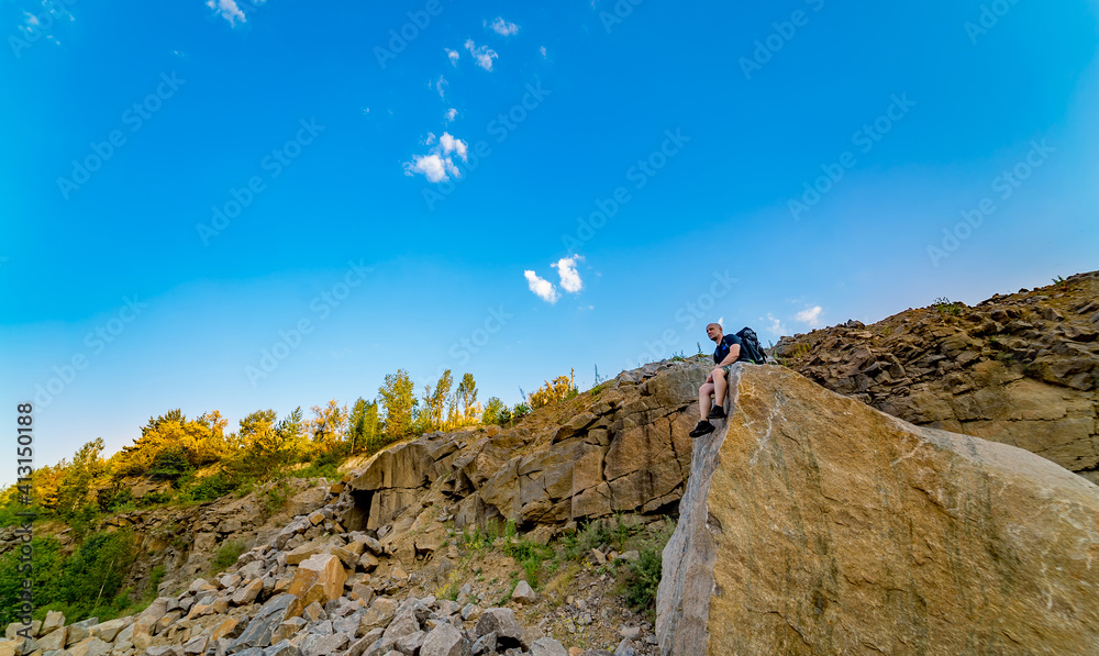 Young man with backpack sitting on a cliffs edge on the top of mountain with gorgeous view. Closeup.