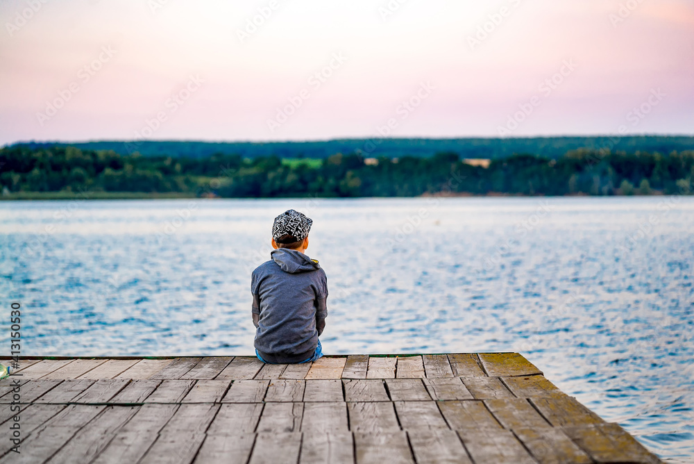 Boy sits on wooden bridge near a pond. Rear view from a back on small boy. Beautiful landscape.