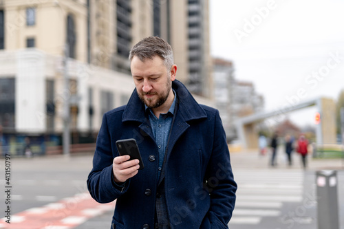 Man walks on a street and looks attentively on a phone. Male in stylish coat with blurred city view background.