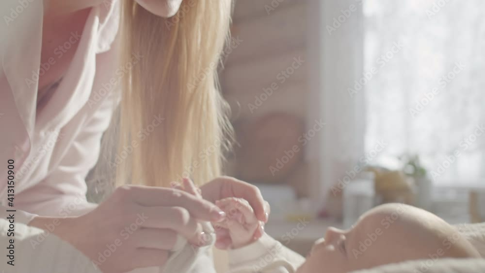 Tilt up shot of beautiful blonde woman standing by bed, holding tiny hands of baby and talking to her at home Tilt up shot of beautiful blonde woman standing by bed, holding tiny hands of baby and talking to her at home