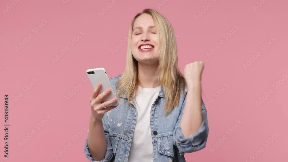 Joyful happy blonde young woman 20s wearing denim jacket isolated on pastel pink color background studio. People lifestyle concept. Using mobile cell phone typing browsing doing winner gesture say yes Joyful happy blonde young woman 20s wearing denim jacket isolated on pastel pink color background studio. People lifestyle concept. Using mobile cell phone typing browsing doing winner gesture say yes