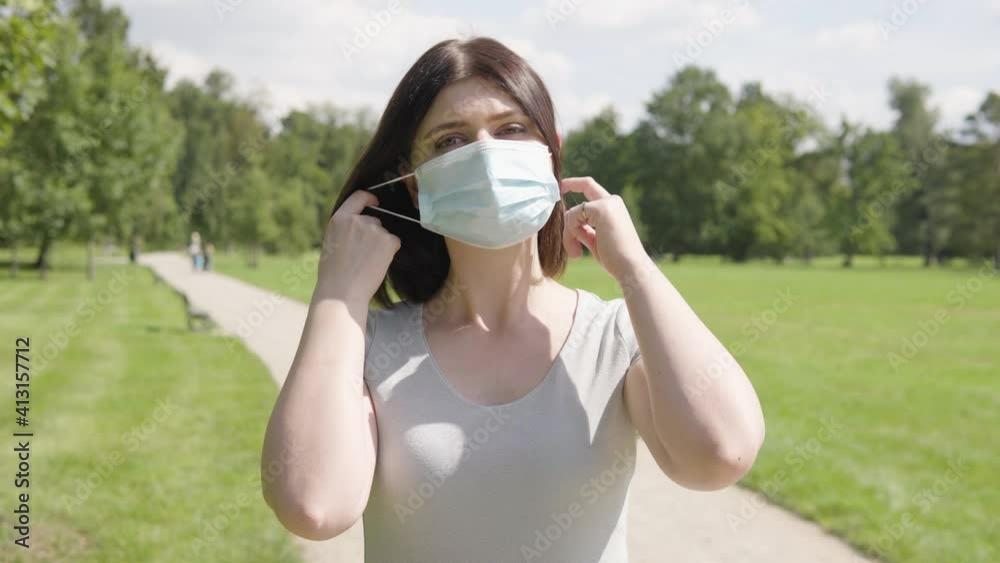 A young Caucasian woman takes off a face mask and looks at the camera on a pathway in a park on a sunny day A young Caucasian woman takes off a face mask and looks at the camera on a pathway in a park on a sunny day