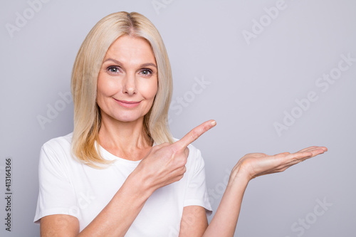 Photo portrait of positive elderly woman pointing finger at holding in hand blank space isolated on grey colored background