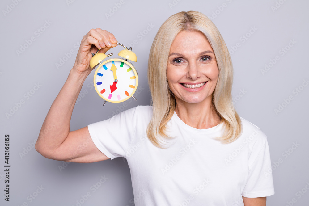 Portrait of charming aged woman toothy smile hand hold classic clock isolated on grey color background Portrait of charming aged woman toothy smile hand hold classic clock isolated on grey color background