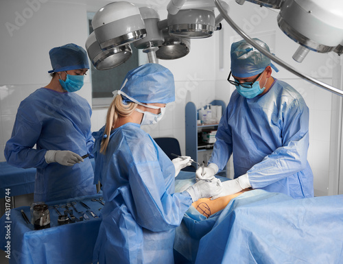 Group of doctors in blue surgical uniforms doing cosmetic surgery in operating room. Surgical team wearing protective face masks, sterile gloves and medical caps. Concept of plastic surgery.