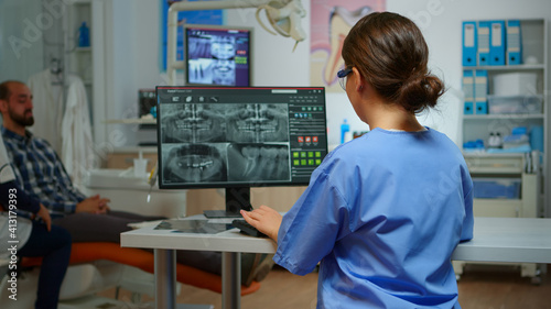 Portrait of smiling nurse in dental office while doctor is working with patient in background. Stomatologist assistant looking on webcam sitting on chair in stomatological clinic.