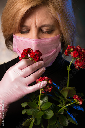 Woman in a mature age in a mask is trying to catch the smell of a flower. Keeps the flowerpot in protective gloves.