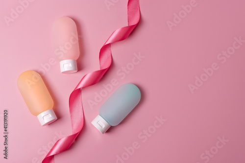 Colorful plastic containers for shampoo, hair balm and shower gel with pink ribbon. Cosmetic products for spa and bath accessories on pink background. Beauty, travel concept, flat lay with copyspace