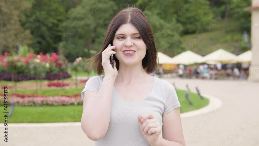 A young Caucasian woman talks on a smartphone with a smile in a city park on a sunny day - people at tables in a restaurant garden in the blurry background A young Caucasian woman talks on a smartphone with a smile in a city park on a sunny day - people at tables in a restaurant garden in the blurry background