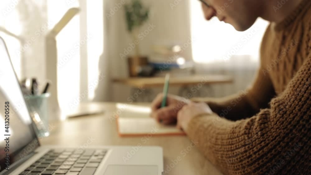 Side view of crop male entrepreneur writing in notebook while planning timetable and sitting at table in home office during remote work 