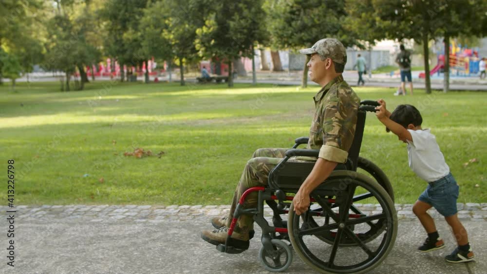 Cute boy helping disabled military dad to wheel heavy wheelchair. Ex soldier walking with son in park. Veteran of war or returning home concept