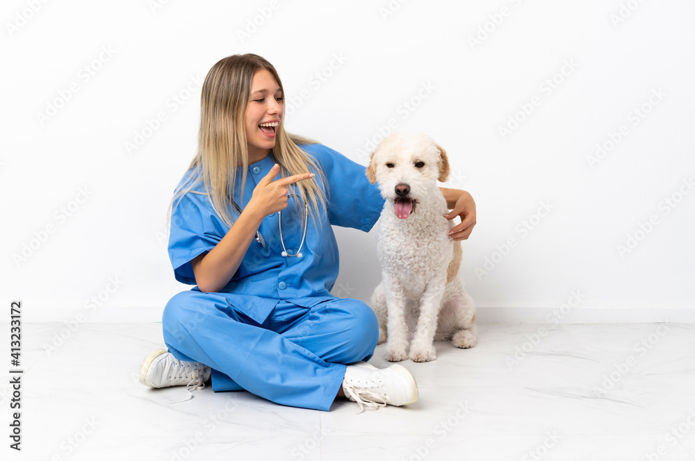 Young veterinarian woman with dog sitting on the floor pointing finger to the side and presenting a product