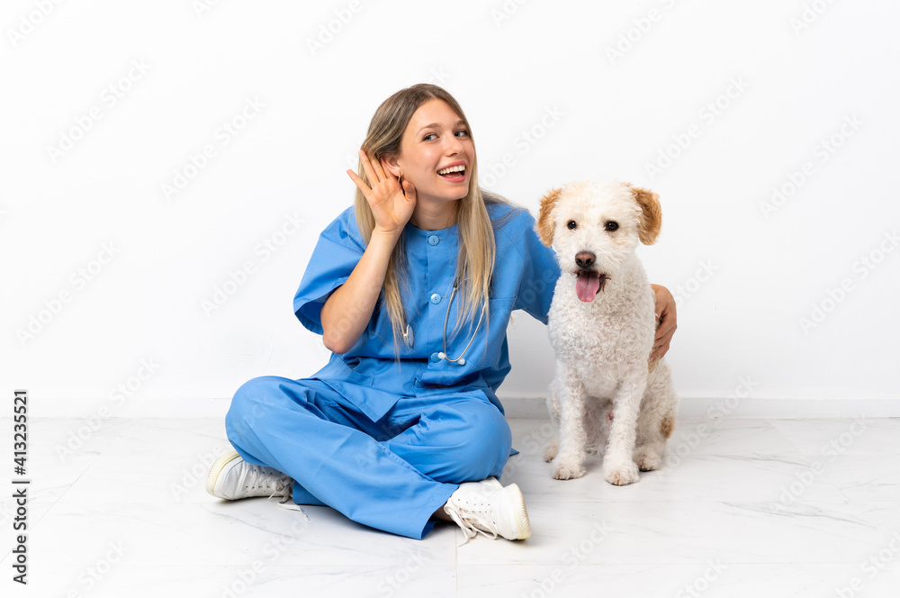 Young veterinarian woman with dog sitting on the floor listening to something by putting hand on the ear
