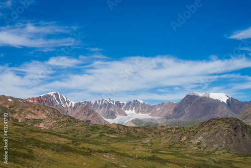 Atmospheric mountain scenery with big beautiful glacier and sharp rocks under blue cloudy sky. Scenic alpine landscape with large glacier tongue. Awesome view from green mountain pass to brown rocks.