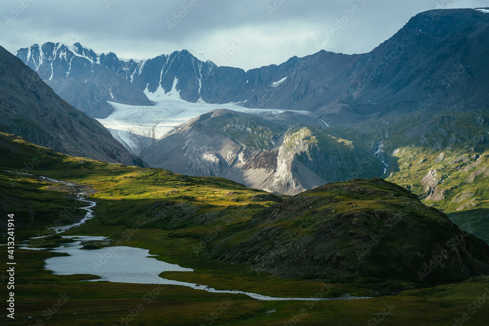 Fototapeta premium Atmospheric alpine landscape with mountain lake in green valley and glacier under cloudy sky. Awesome highland scenery with beautiful glacial lake among sunlit hills and rocks against mountain range.