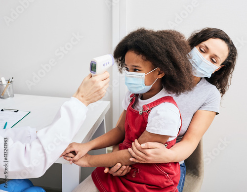 Pediatrician checking body temperature of a child using a non-contact thermometer. African American girl with her mom wearing protective masks at the doctor s consultation