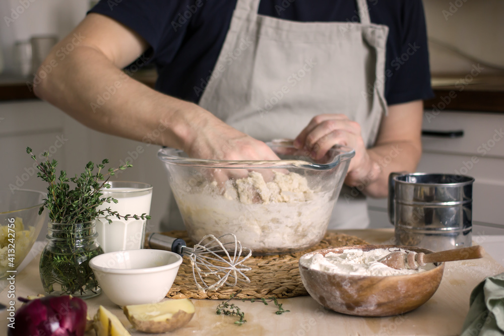 A faceless man in an apron in his kitchen at the kitchen table kneads bread dough in a glass bowl with his hands. Home authentic hobby, home baker. Baking bread with your own hands